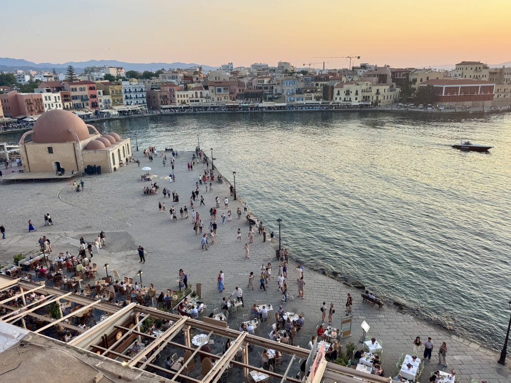 The old town of Shania, Greece, at sunset. A calm harbor with a single boat in it, and a yellow, orange, and purple sky over the short buildings. Many people are out and about along the harbor's edge.
