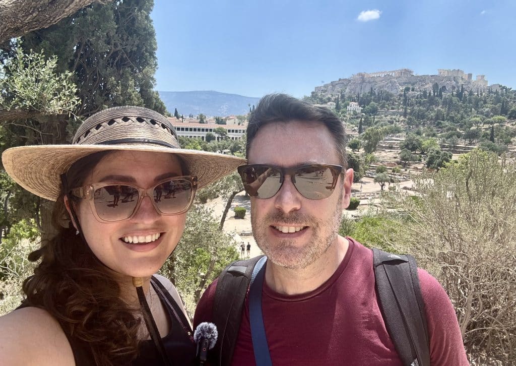Kate and Charlie take a smiling selfie in Greece with the Acropolis rising up on a hill behind them. They both wear sunglasses and Kate wears a wide-brimmed straw hat.
