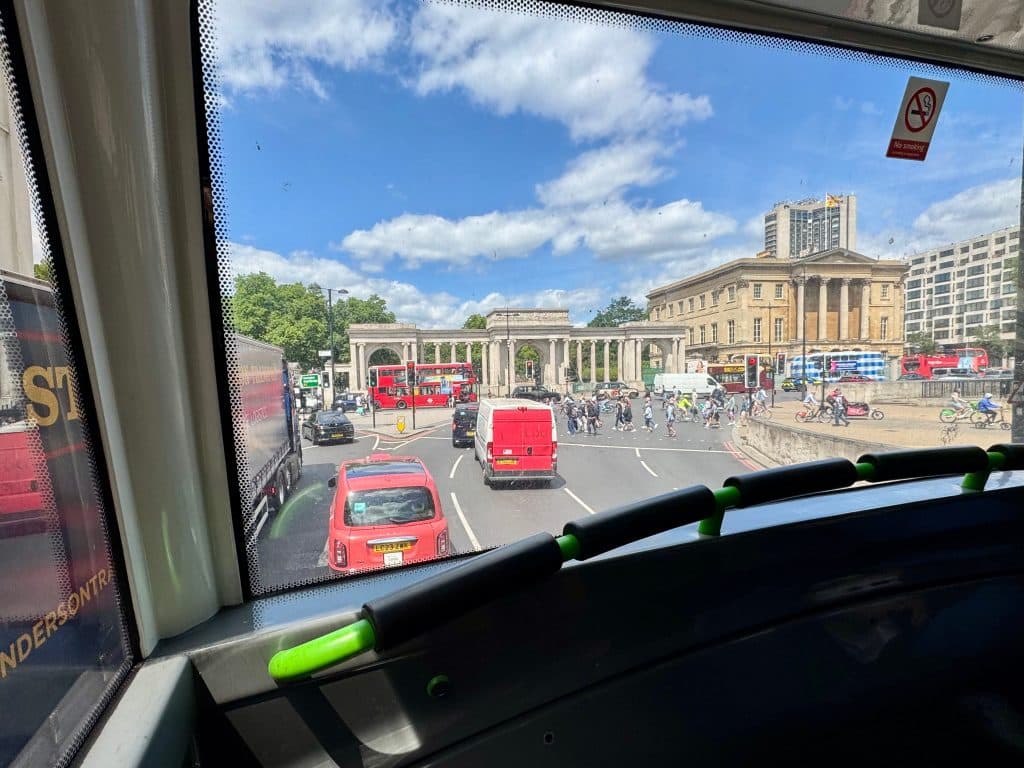 A view of London through the window of a Double Decker Bus, lots of traffic going around a circle and some old-fashioned church-like buildings.