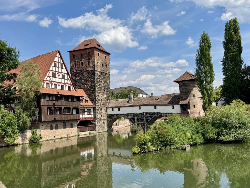 Beautiful half-timbered buildings and a stone tower in Germany, set on a still river underneath a cloud-streaked sky.