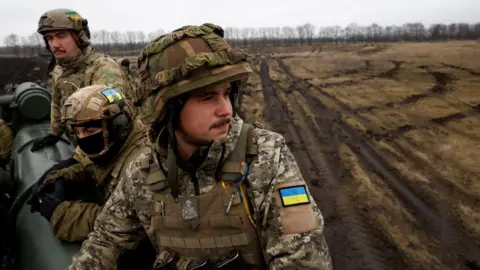 Reuters Ukrainian soldiers in full battle dress sit on a cannon in a muddy field.  