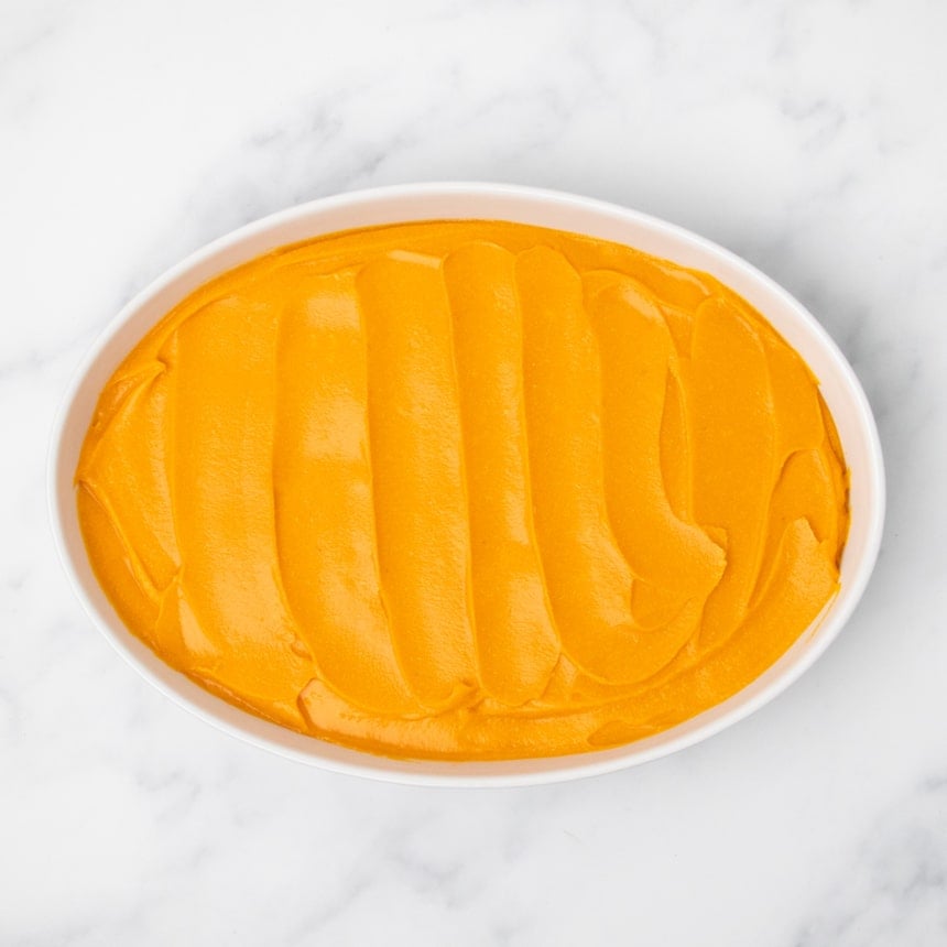 A white oval baking dish filled with evenly spread orange sweet potato filling on a white marble surface.
