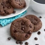 Two keto chocolate breakfast cookies on a gray table with a cup of coffee and more cookies on a plate in the background.