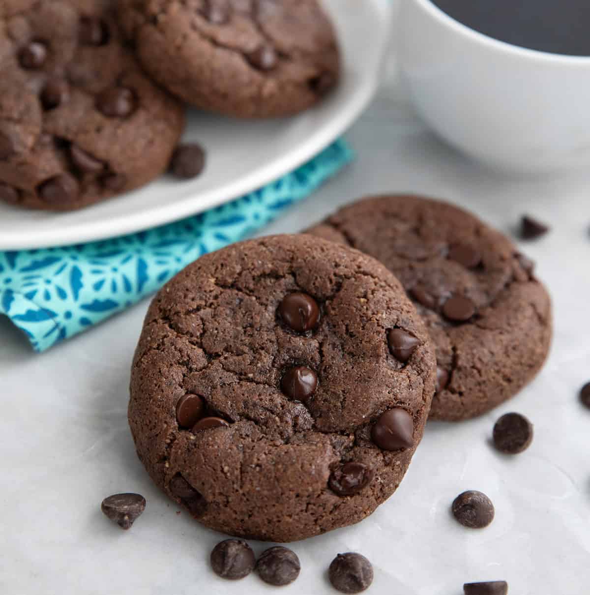 Two keto chocolate breakfast cookies on a gray table with a cup of coffee and more cookies on a plate in the background.