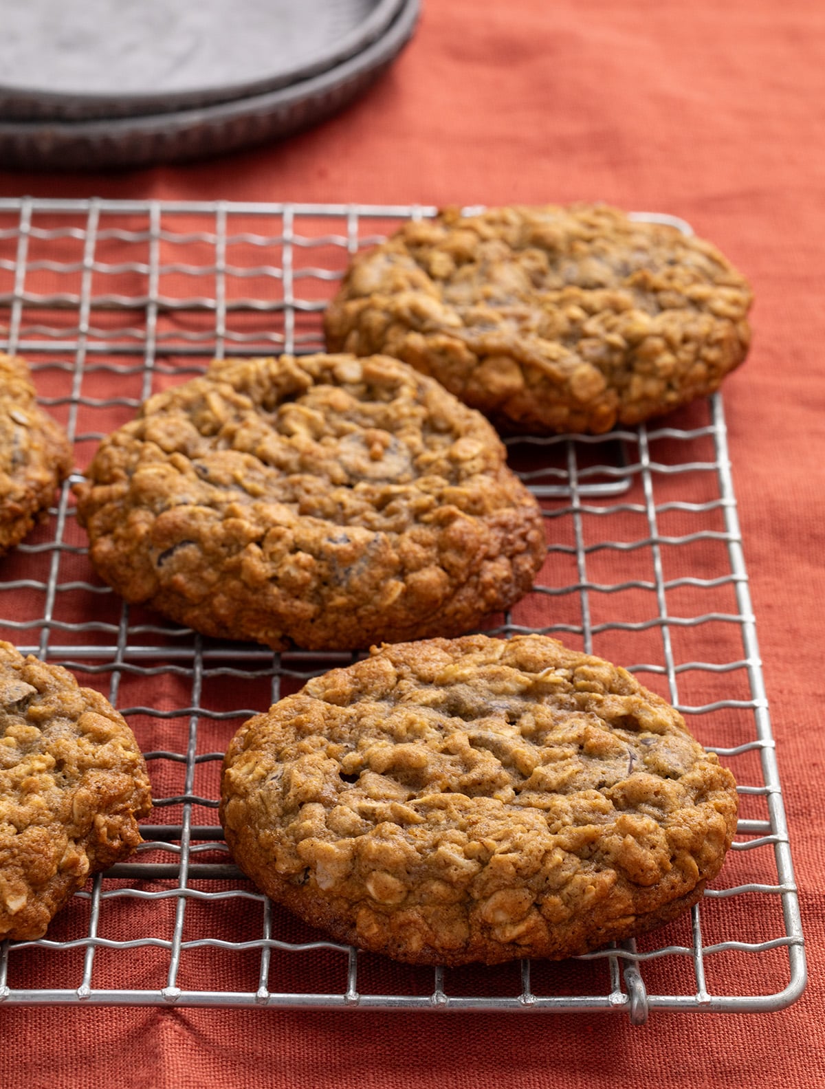 Five oatmeal cookies on a metal cooling rack, only 3 completely visible in the frame, with stacked gray plates in the background, all on an orange fabric surface.