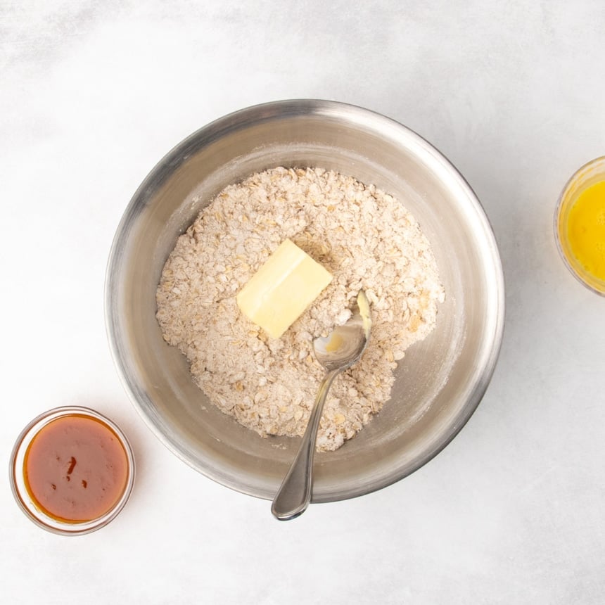 A metal mixing bowl filled with a mixture of pale brown dry ingredients and a piece of butter on top, with a metal mixing spoon resting inside the bowl; two small bowls, one containing orange puree and one with yellow beaten egg, are nearby.