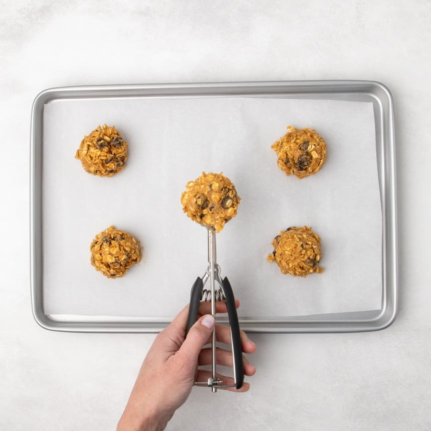 A hand holding a black-handled cookie scoop with raw cookie dough above a parchment-lined baking sheet with four other portions of raw rust-colored oatmeal cookie dough spaced apart.