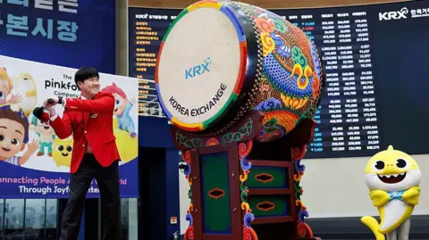 Getty Images Kim Min-seok, chief executive officer of Pinkfong, strikes the drum during the firm's listing ceremony at the Korea Exchange in Seoul, South Korea, on 18 November 2025. He is dressed in a red suit as he swings a baton at a multi-coloured drum while a yellow Baby Shark mascot standing on the right of the image cheers him on.