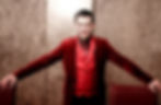 Melbourne magician Richard Vegas dressed in a red velvet jacket, a red satin vest and a red tie, stands with his arms resting on the back of 2 chairs.