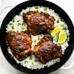 Jerk chicken thighs served on a bed of fluffy white rice in a black skillet, topped with chopped green onions and lime wedges on the side.