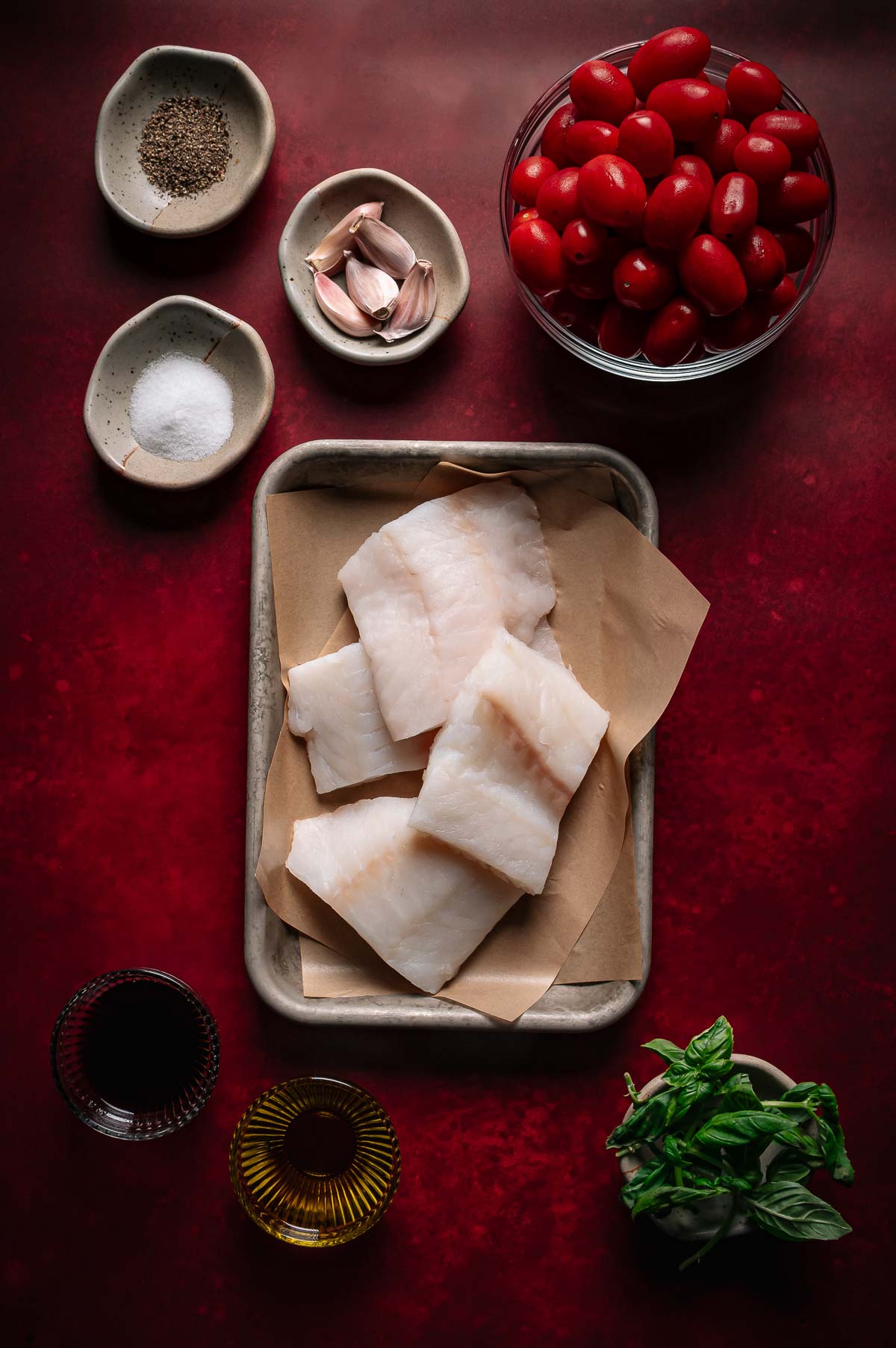 Raw cod fillets on parchment with bowls of grape tomatoes, garlic, basil, salt, pepper, olive oil, and balsamic vinegar, all arranged on a red surface.
