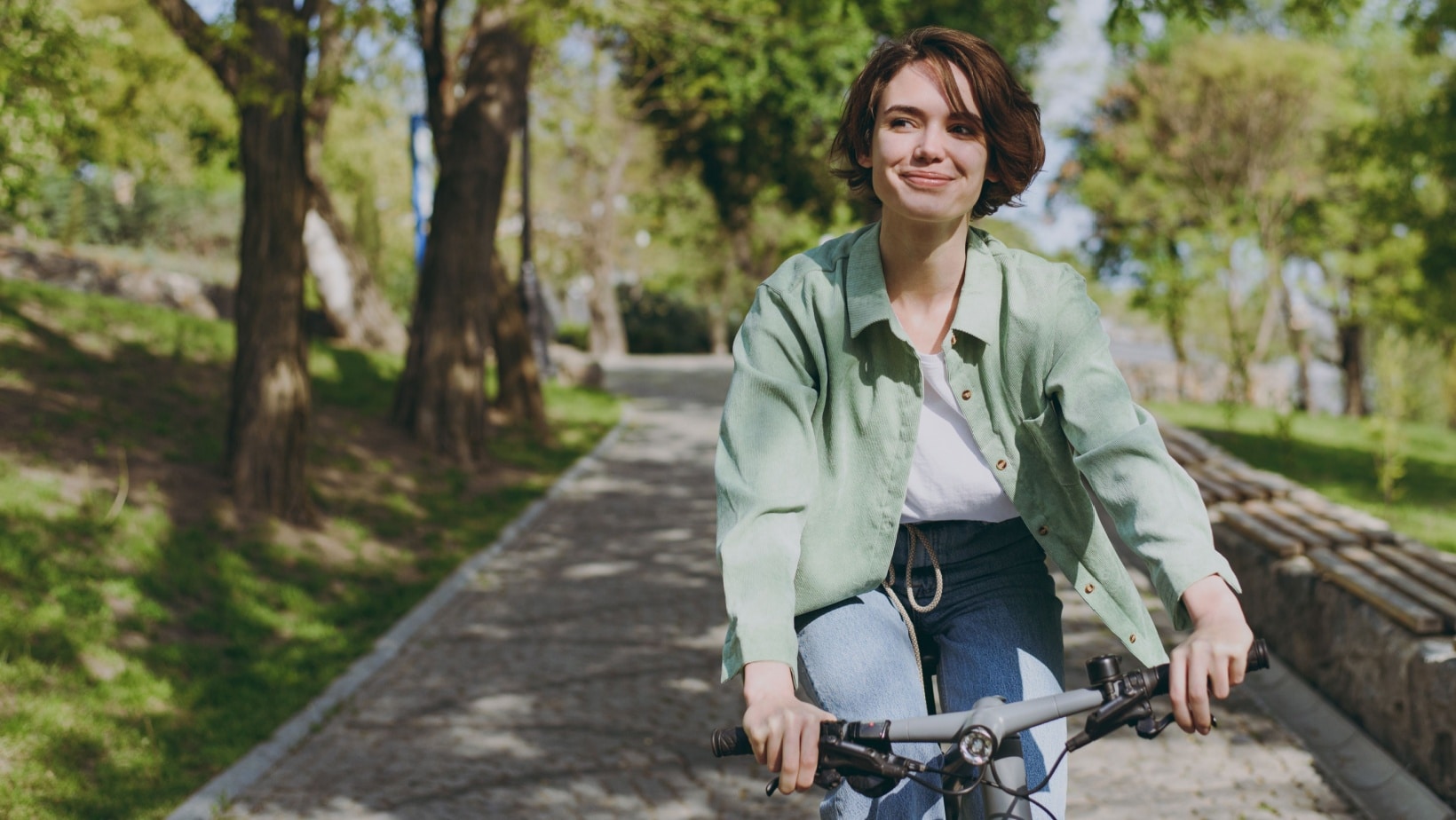 Young pensive dreamful happy woman 20s wearing casual green jacket jeans riding bicycle bike on sidewalk in city spring park outdoors, look aside. People active urban healthy lifestyle cycling concept