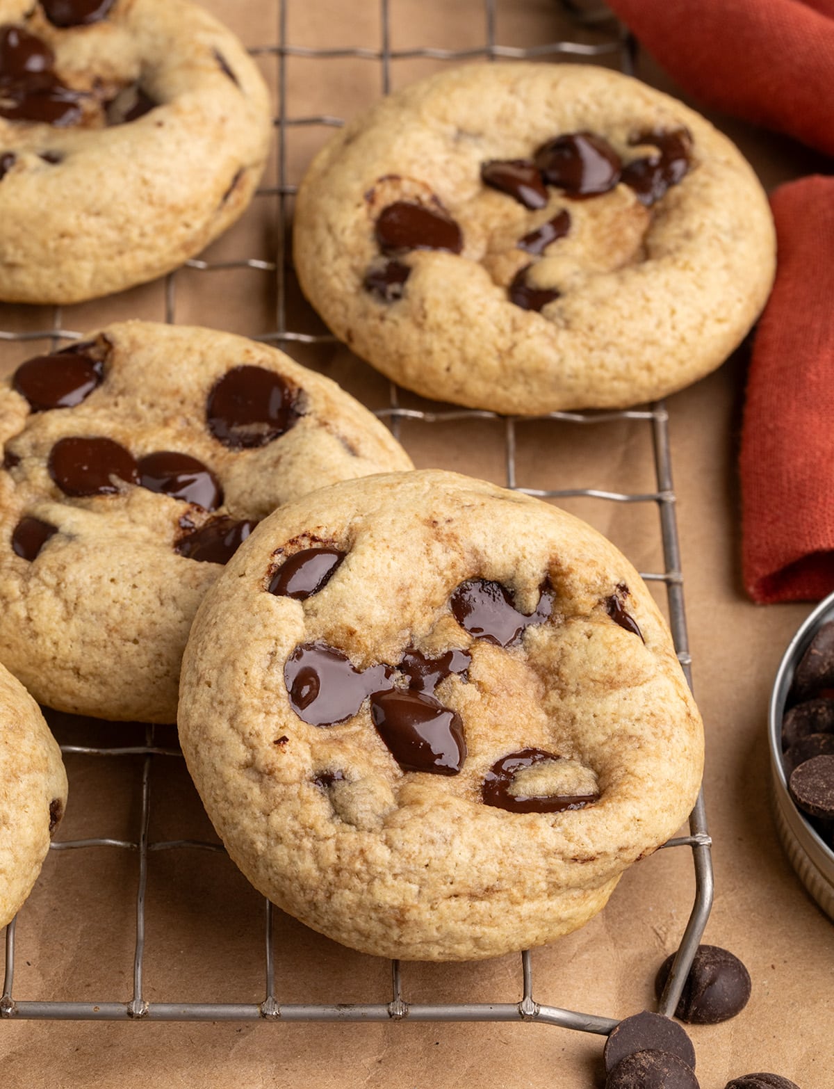 A close-up of chocolate chip cookies on a wire rack, with chocolate chips and a red cloth nearby.