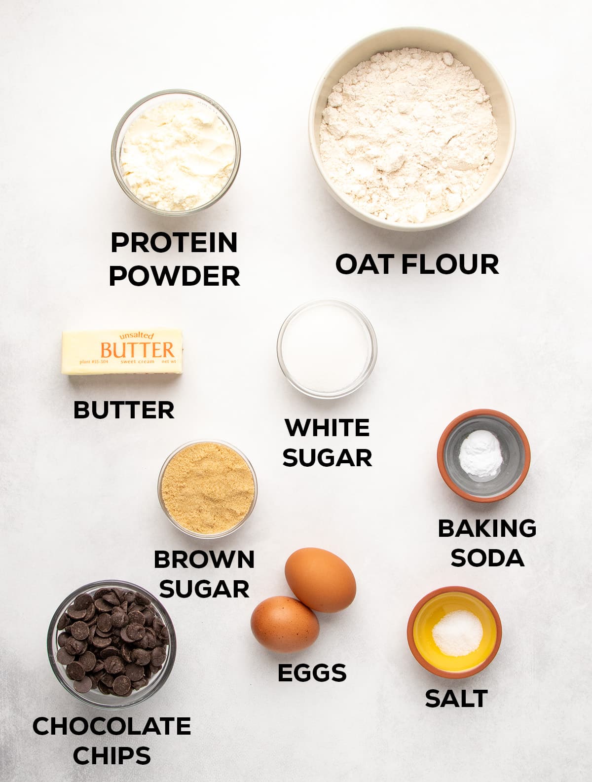 Ingredients arranged on a light surface with text labels: protein powder, oat flour, butter, white sugar, baking soda, brown sugar, eggs, salt, and chocolate chips.