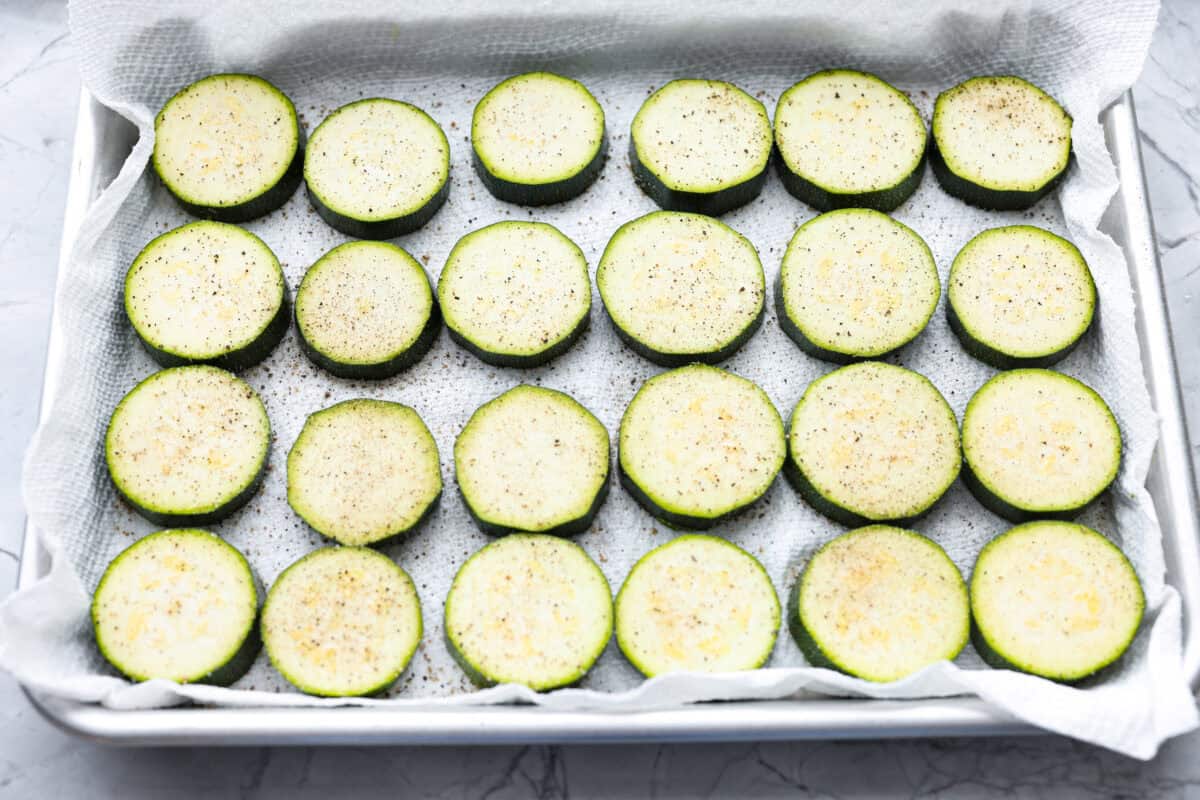 Zucchini slices on parchment paper on a baking sheet with salt and pepper sprinkled over them 
