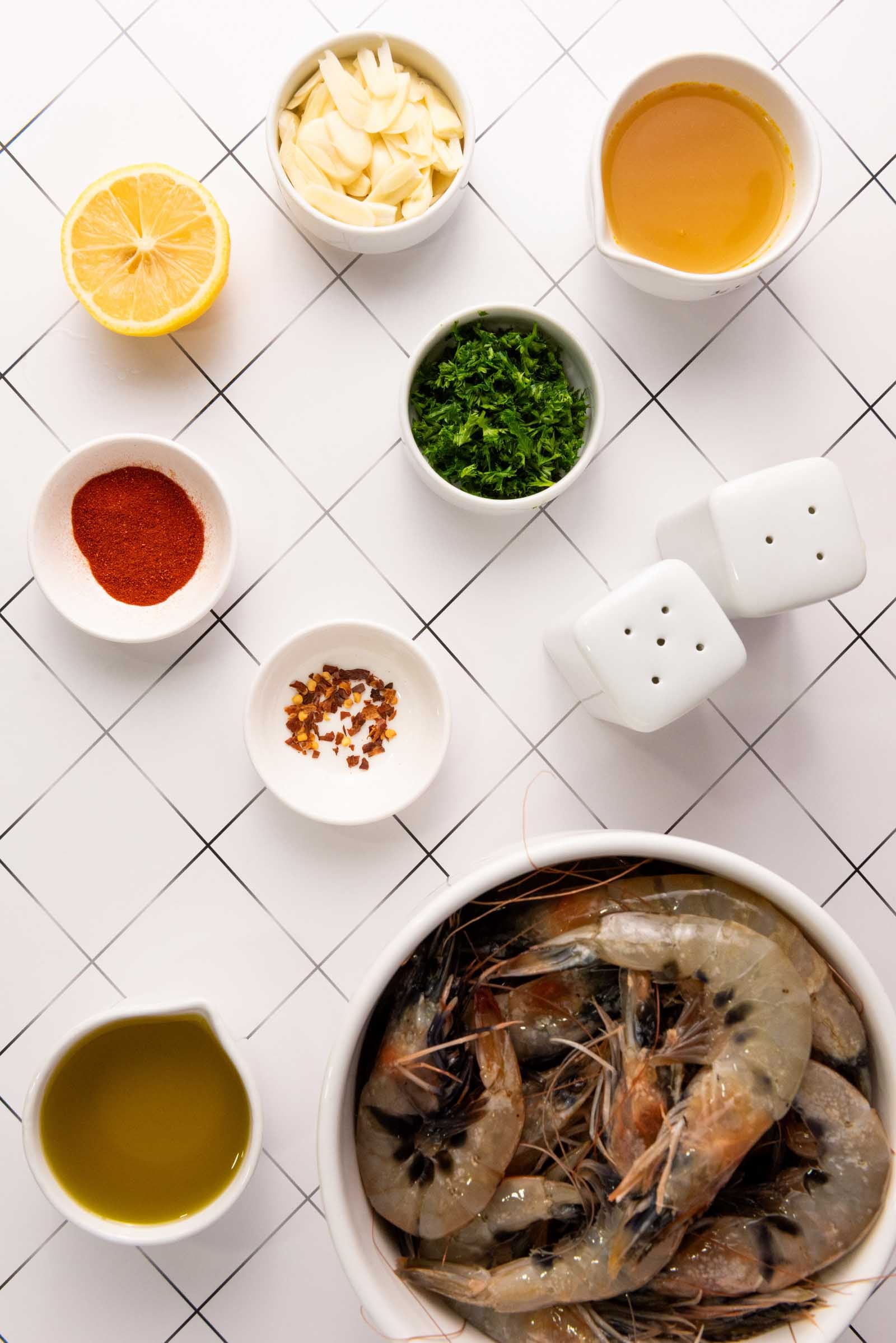 Raw shrimp in a bowl with small dishes of sliced garlic, olive oil, chopped parsley, lemon, paprika, red pepper flakes, and broth on a white tiled surface.