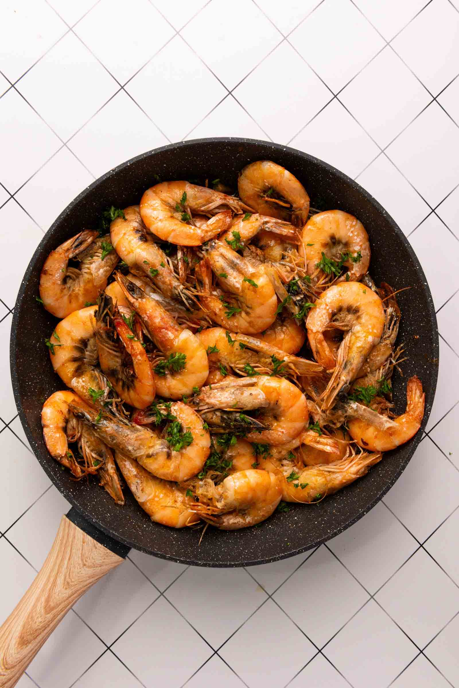 Cooked shrimp with herbs in a black frying pan on a white tiled surface, top-down view.