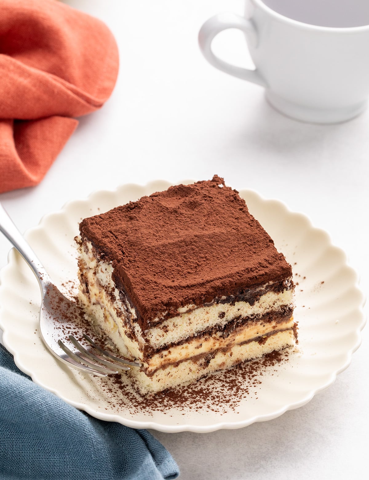 Square slice of tiramisu on a scalloped white plate with cocoa powder on top, a fork beside it, a blue cloth, an orange cloth, and a white cup of coffee nearby.