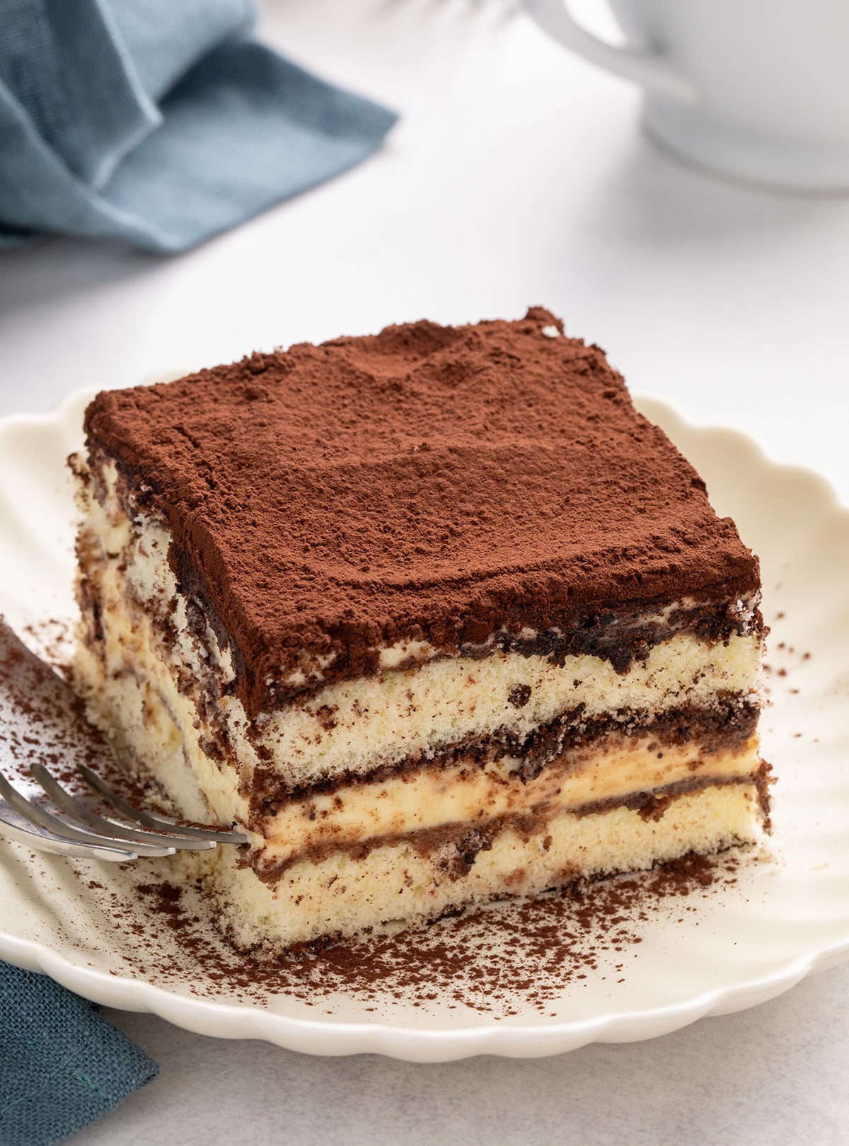 Close-up of a square slice of tiramisu on a scalloped white plate with visible cream layers, cookie layers, and cocoa powder on top.