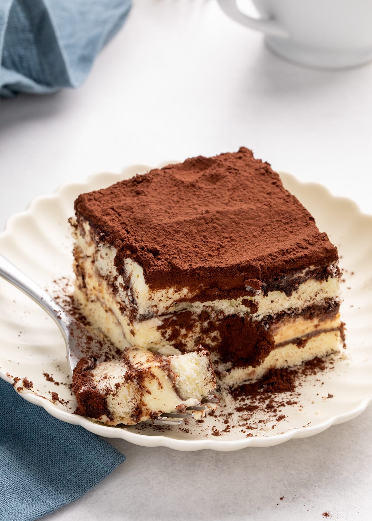 Square slice of tiramisu on a scalloped white plate with a forkful taken from the front.