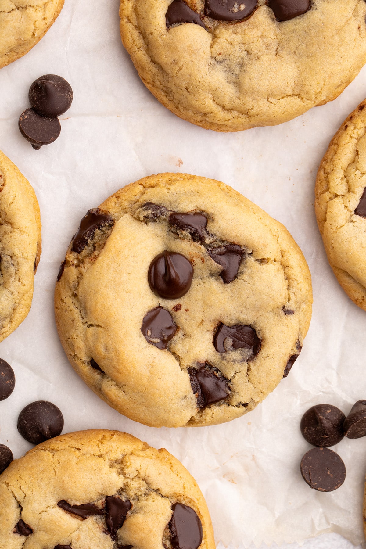 Close-up overhead image of chocolate chip cookies and loose chocolate chips on parchment paper.