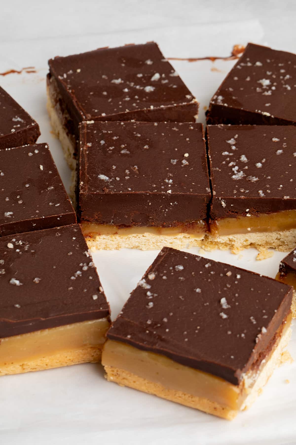 Close-up of layered shortbread squares with chocolate, caramel, and shortbread layers on parchment paper.