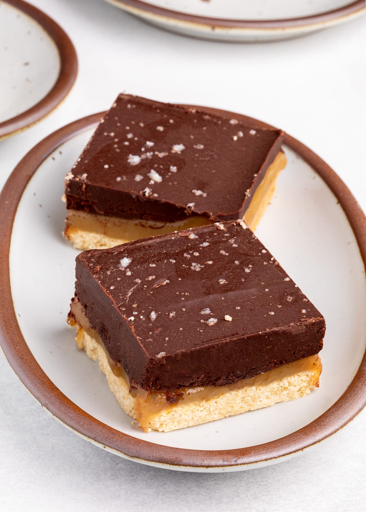 Two layered chocolate, caramel, and shortbread squares on a small oval serving plate with a brown rim.