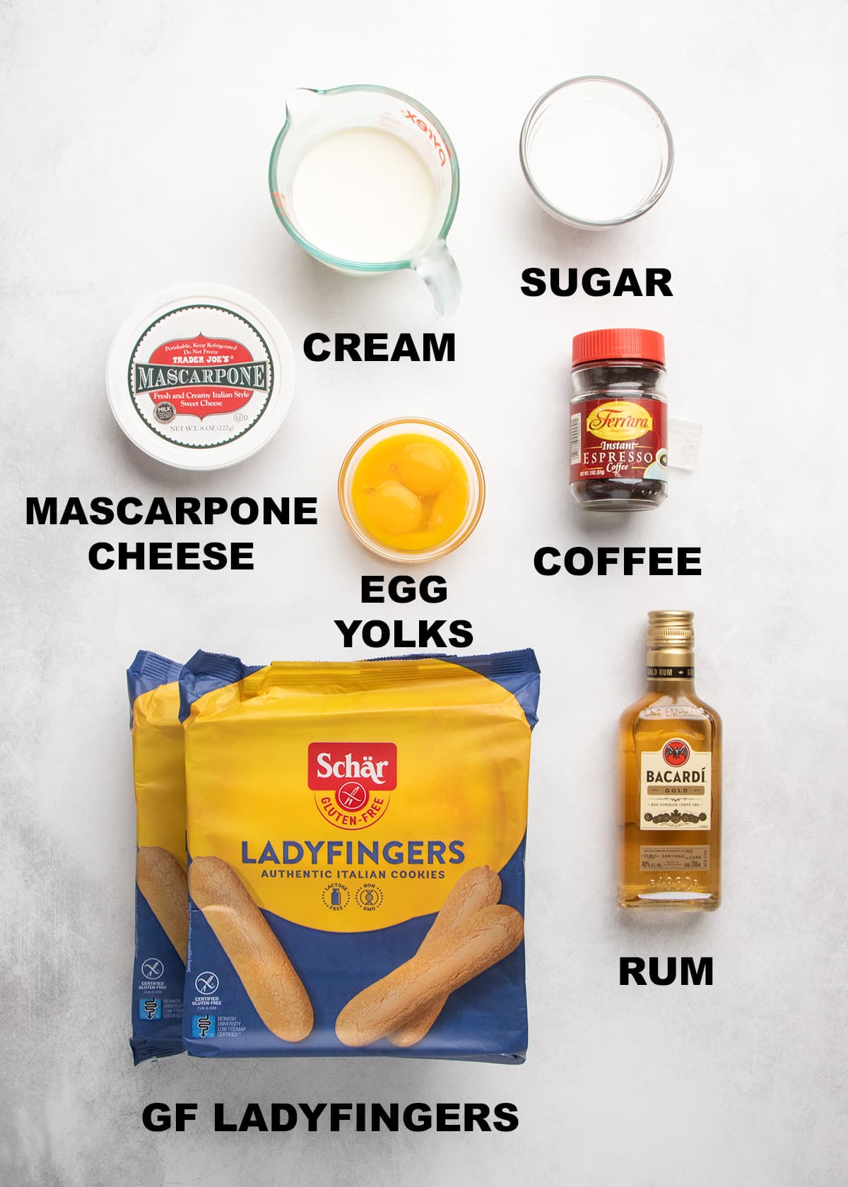 Labeled ingredients for gluten free tiramisu arranged on a light surface, including mascarpone cheese, cream, sugar, egg yolks, coffee, rum, and gluten free ladyfingers.