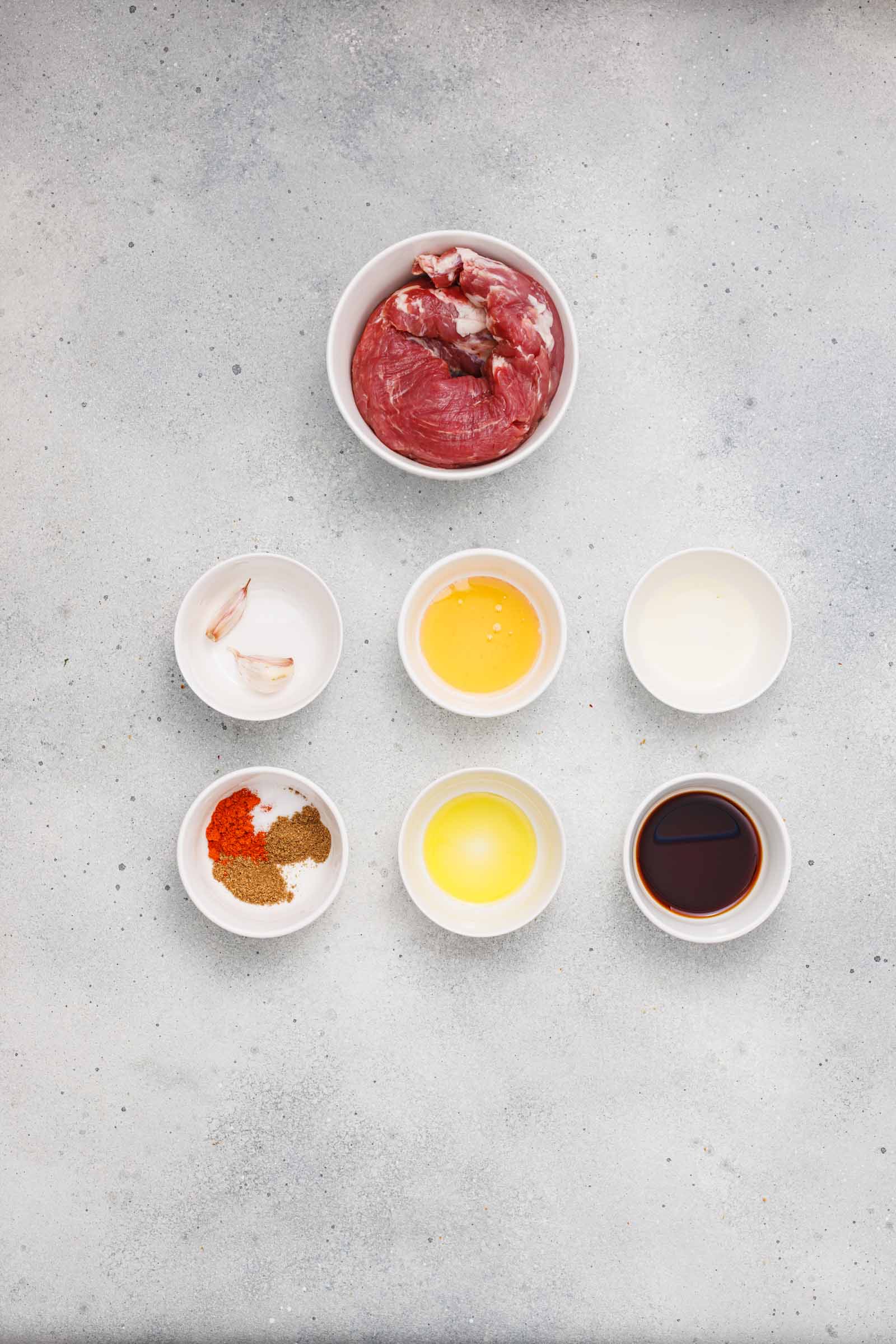 Seven small bowls containing raw beef, garlic cloves, oil, vinegar, spices, lemon juice, and soy sauce arranged on a light gray countertop.
