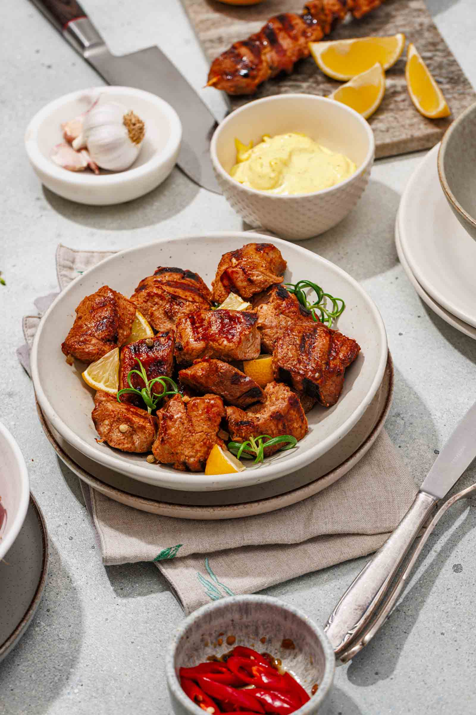 A plate of grilled pork pieces garnished with green onions and lemon wedges, surrounded by garlic, sauce, lemon slices, and sliced red chili on a light tabletop.