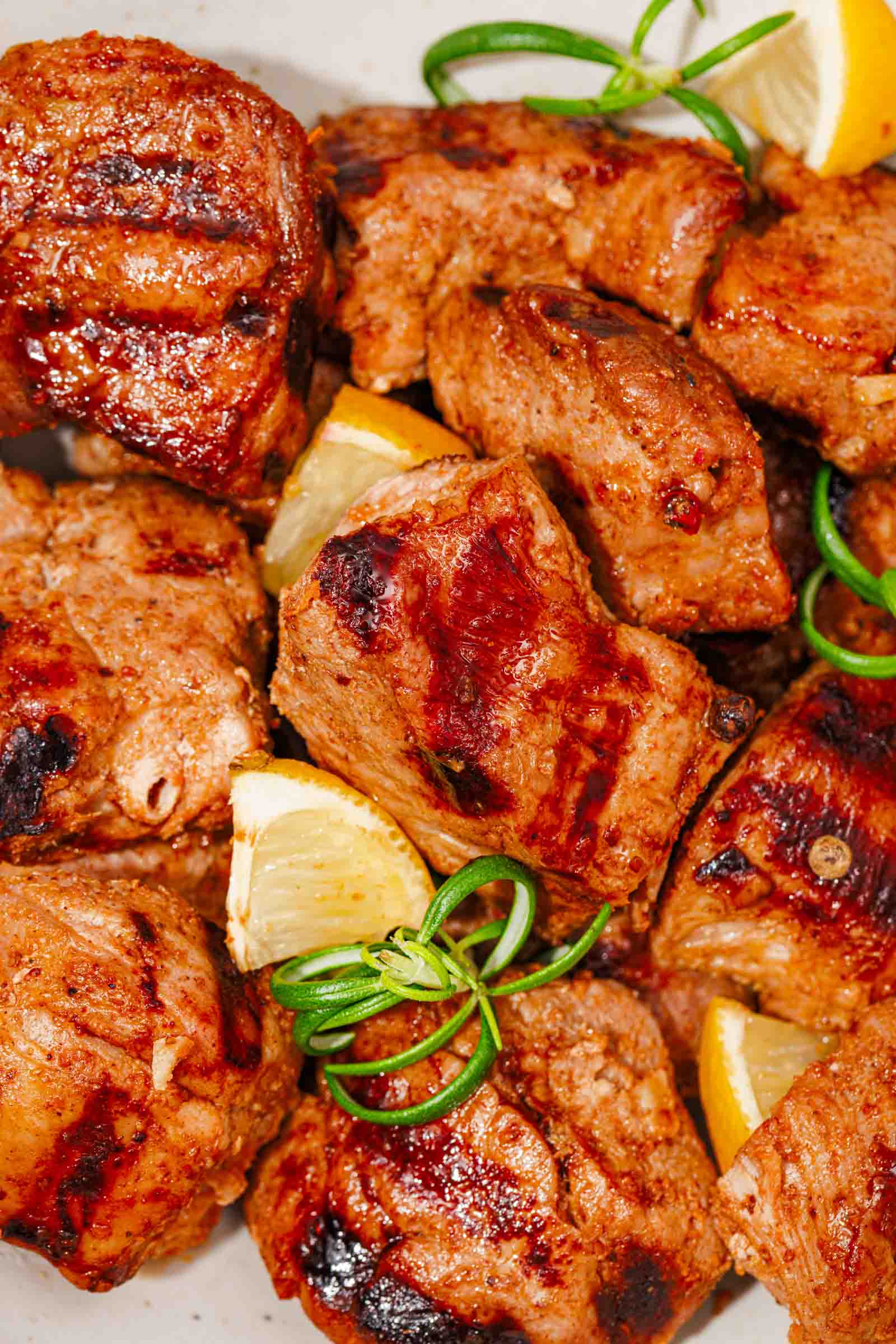 Grilled marinated pork chunks served with lemon wedges and garnished with green herbs on a white plate.