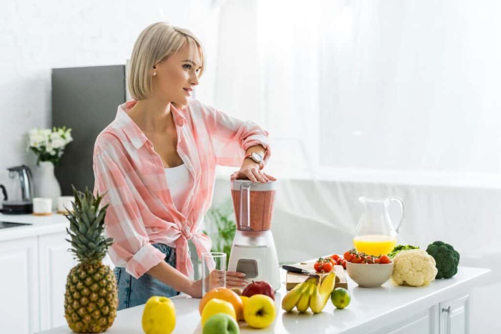 Woman making a smoothie with fruit and vegetables in a bright kitchen.