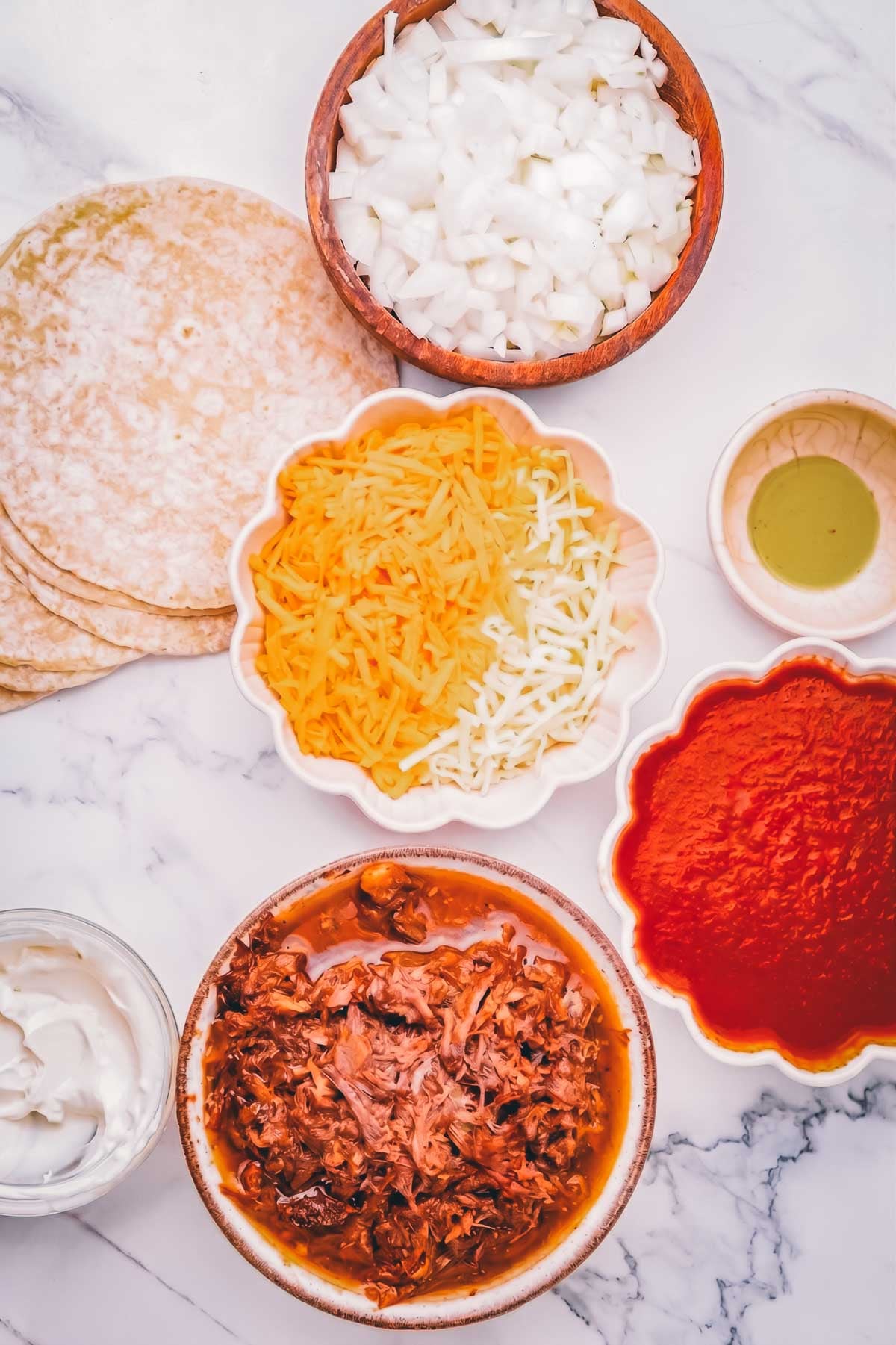Overhead view of shredded mexican beef, grated cheese, chopped onions, tomato sauce, sour cream, corn tortillas, and oil in bowls on a marble surface.