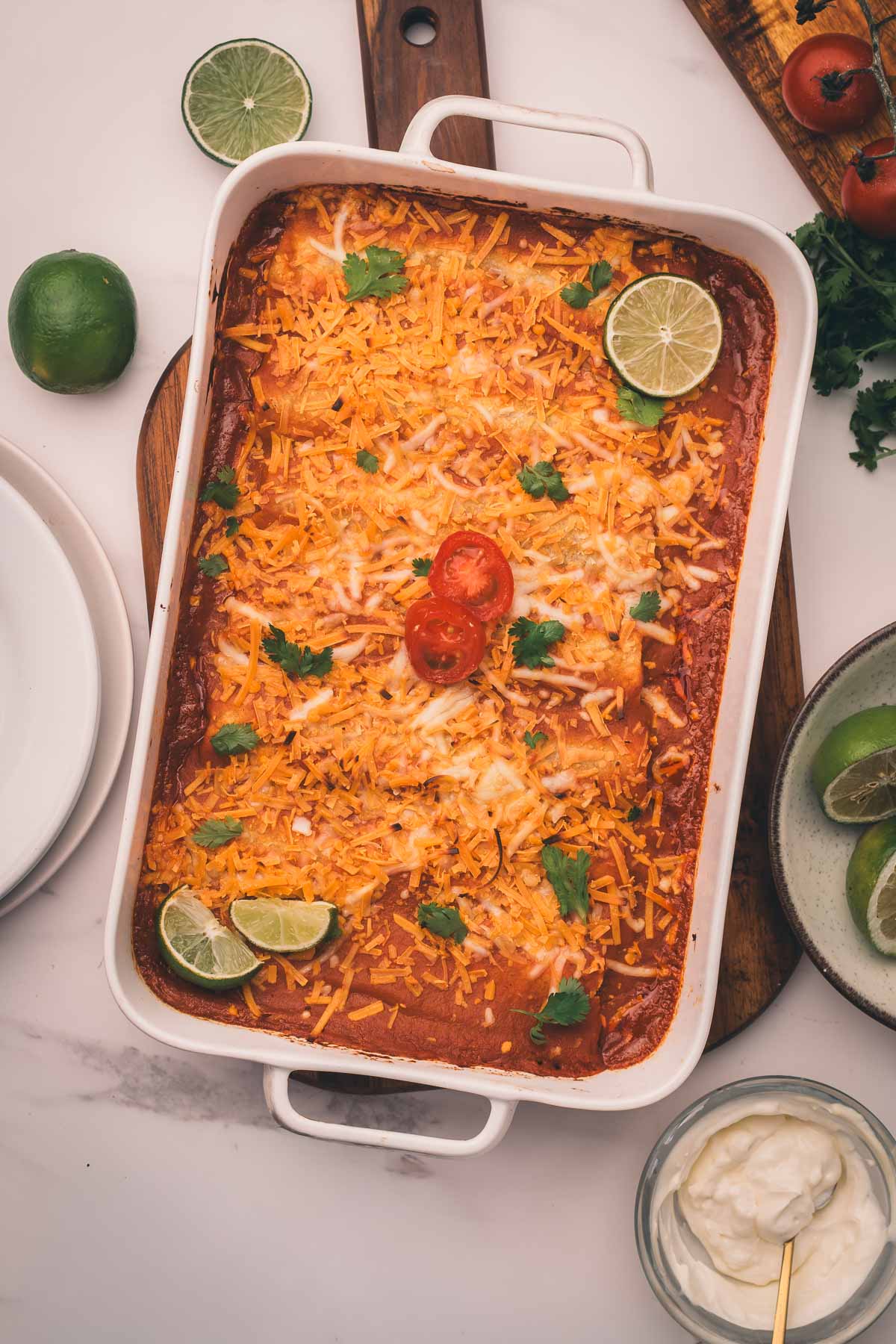 A casserole dish filled with baked enchiladas, topped with melted cheese, cilantro, lime wedges, and sliced cherry tomatoes. Plates, limes, and a bowl of sour cream are nearby.