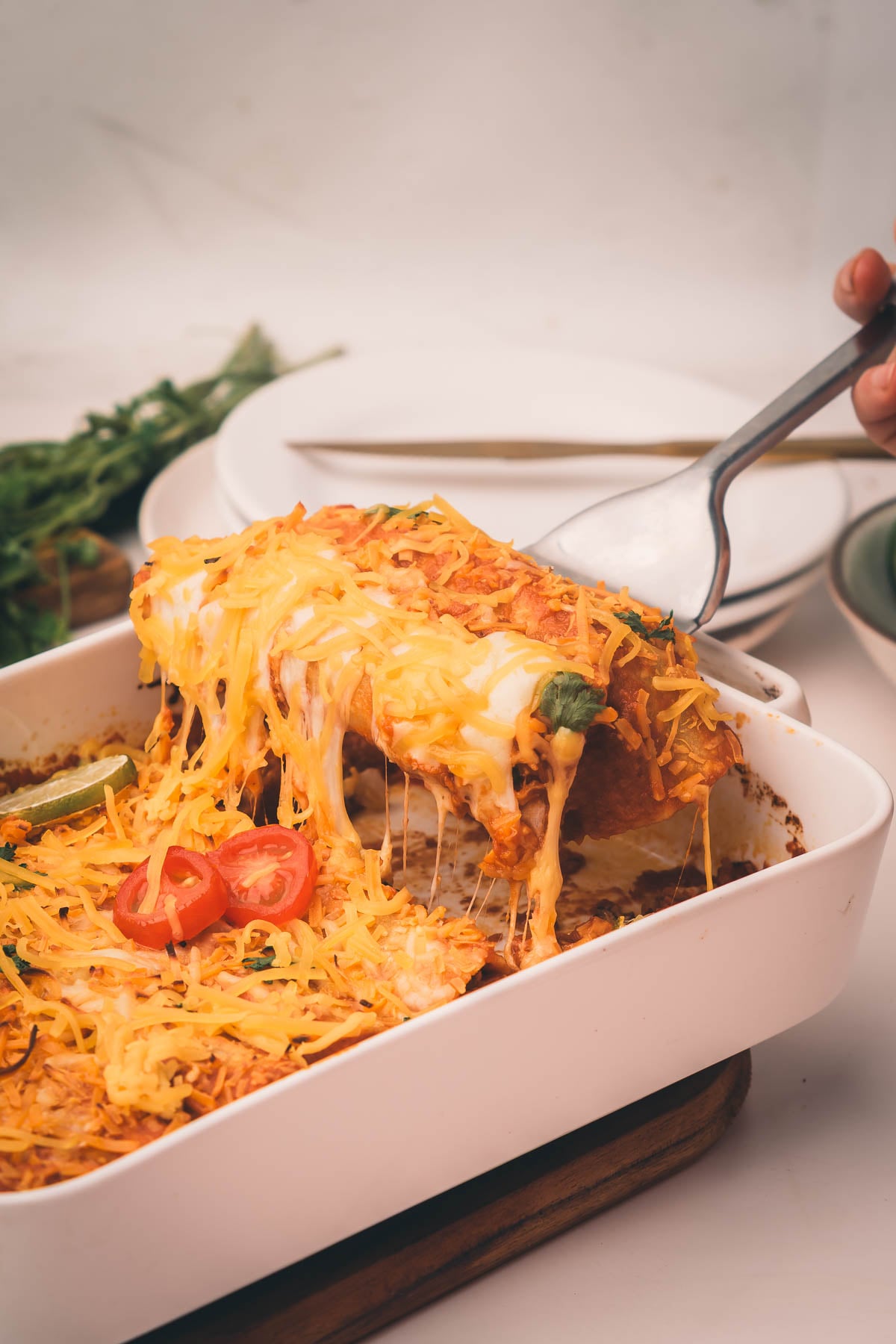 A hand lifts a cheesy shredded beef enchilada from a white baking dish filled with baked enchiladas, topped with melted cheese and sliced cherry tomatoes. Plates and greens are in the background.