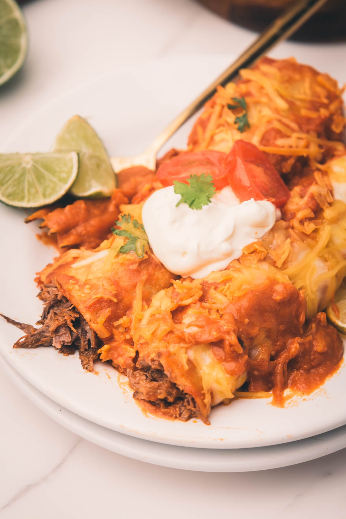 A plate of shredded beef enchiladas topped with melted cheese, tomato sauce, sour cream, cilantro, and lime wedges, served on a white plate.