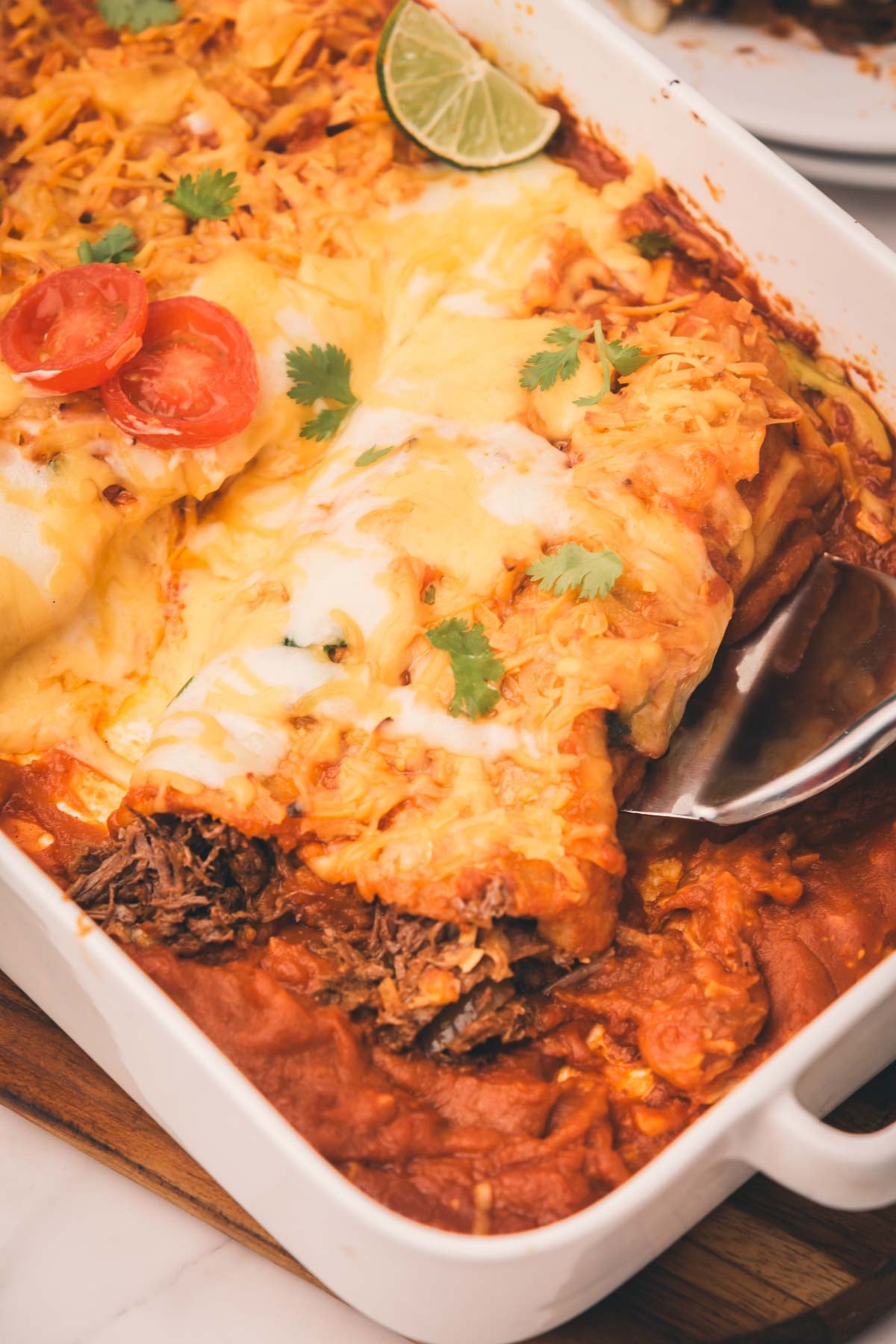 A baking dish of cheesy shredded beef enchiladas topped with melted cheese, tomato slices, cilantro, and a lime wedge. A serving spoon is lifting one enchilada from the dish.
