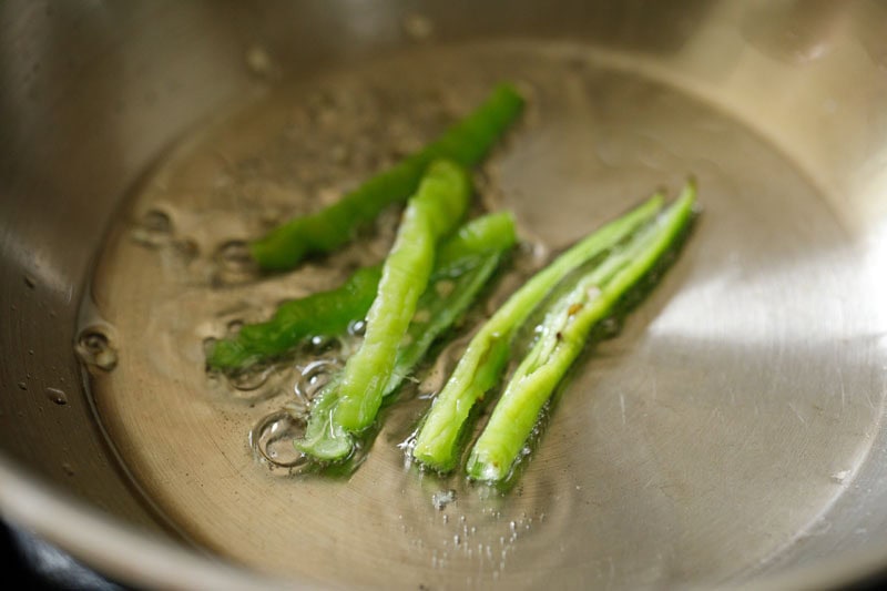 Slit green chillies sizzling in oil in pan.
