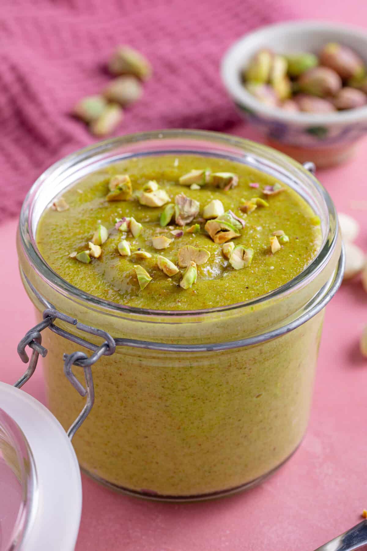 A jar of homemade pistachio cream with a pink backdrop, with crushed pistachio sprinkled on top.