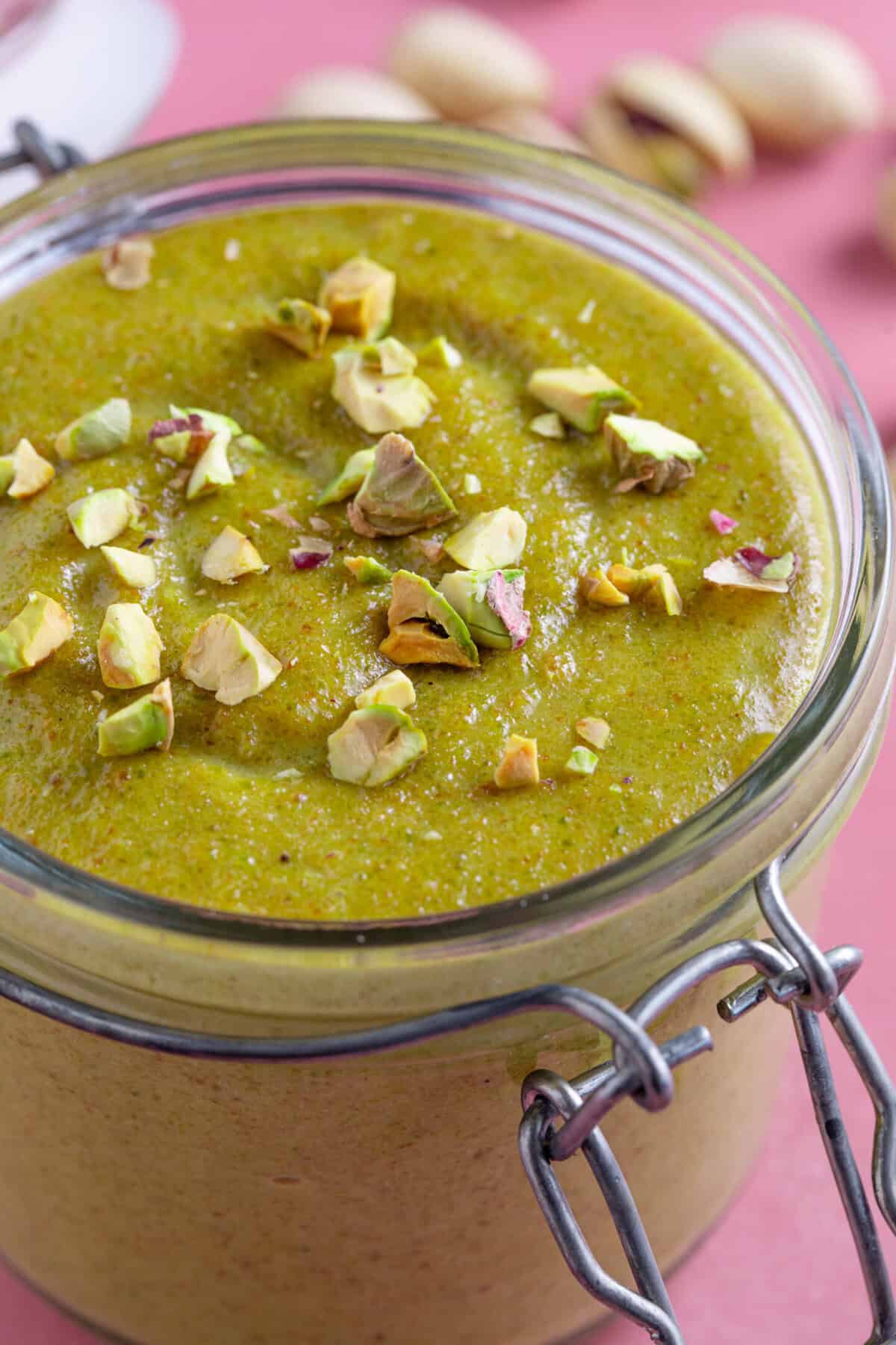 Close up shot of a jar of homemade pistachio cream with a pink backdrop, with crushed pistachio sprinkled on top.
