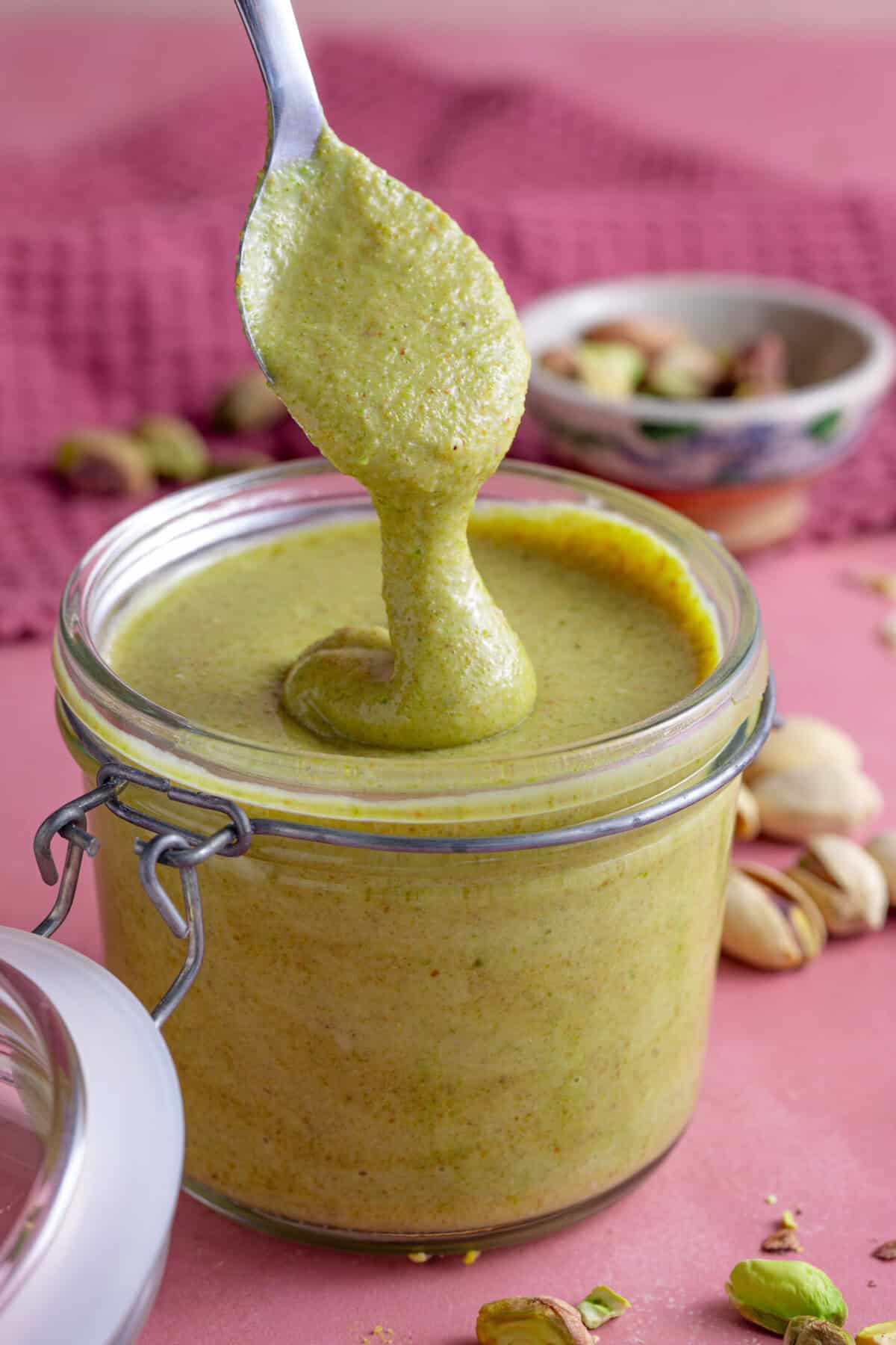 A spoon drizzling smooth pistachio paste into a glass mason jar.