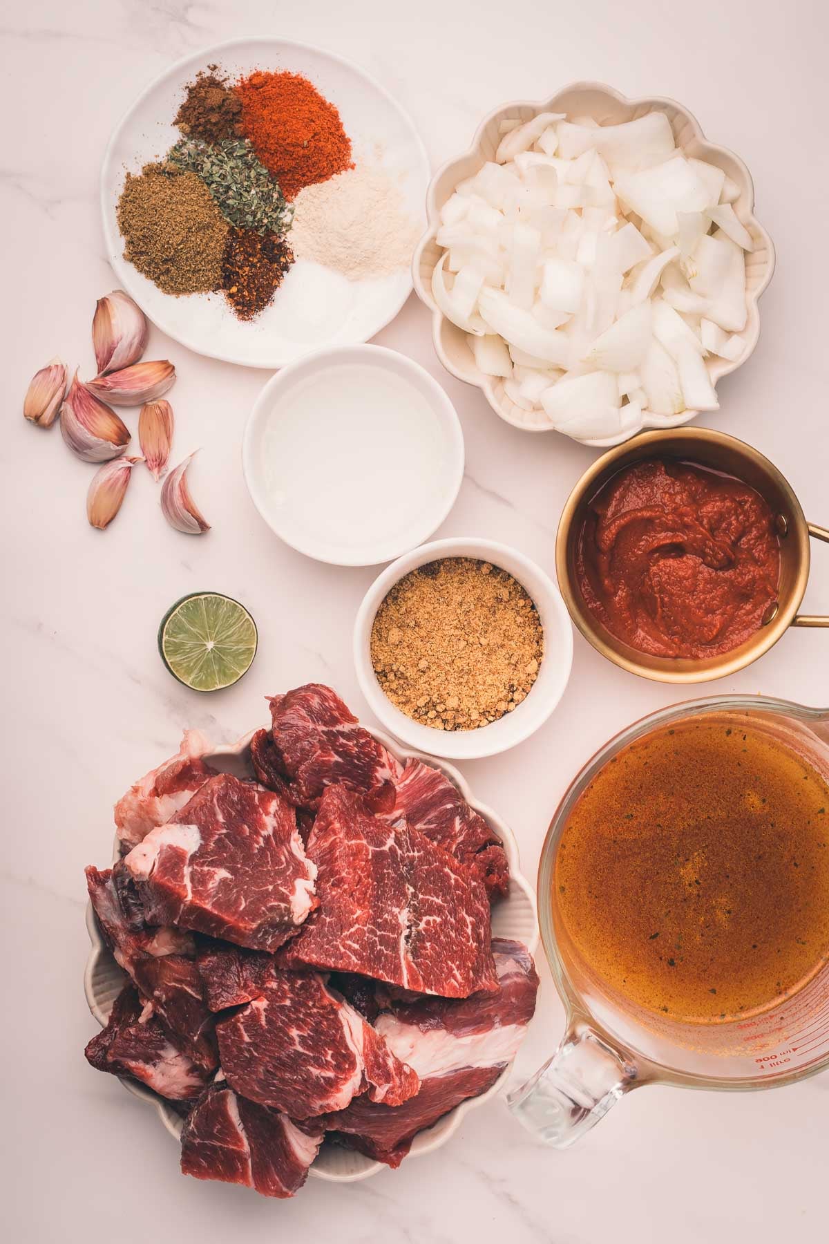 Chunks of raw beef, chopped onions, garlic cloves, a halved lime, spices, tomato paste, broth, brown sugar, and a small bowl of liquid arranged on a white surface.
