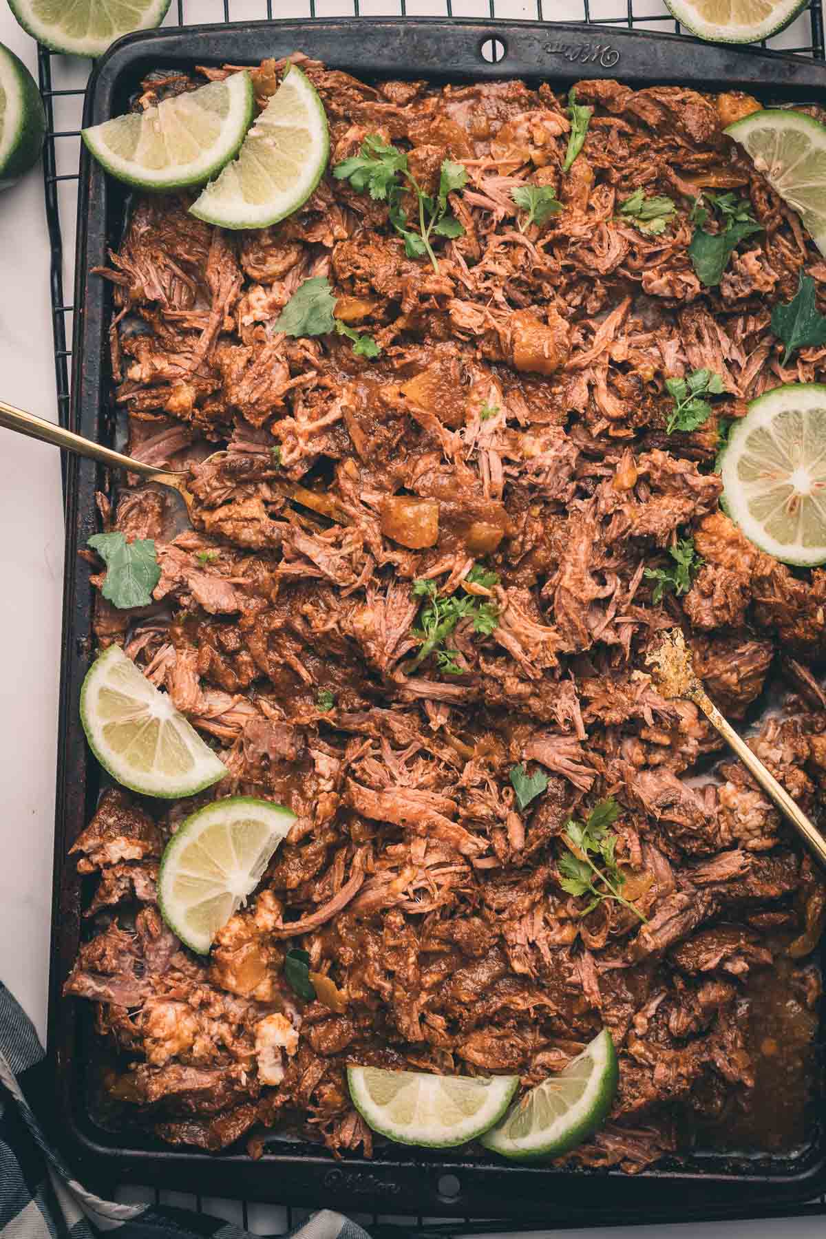 Shredded Mexican beef garnished with cilantro and lime slices on a baking tray, with serving utensils visible.