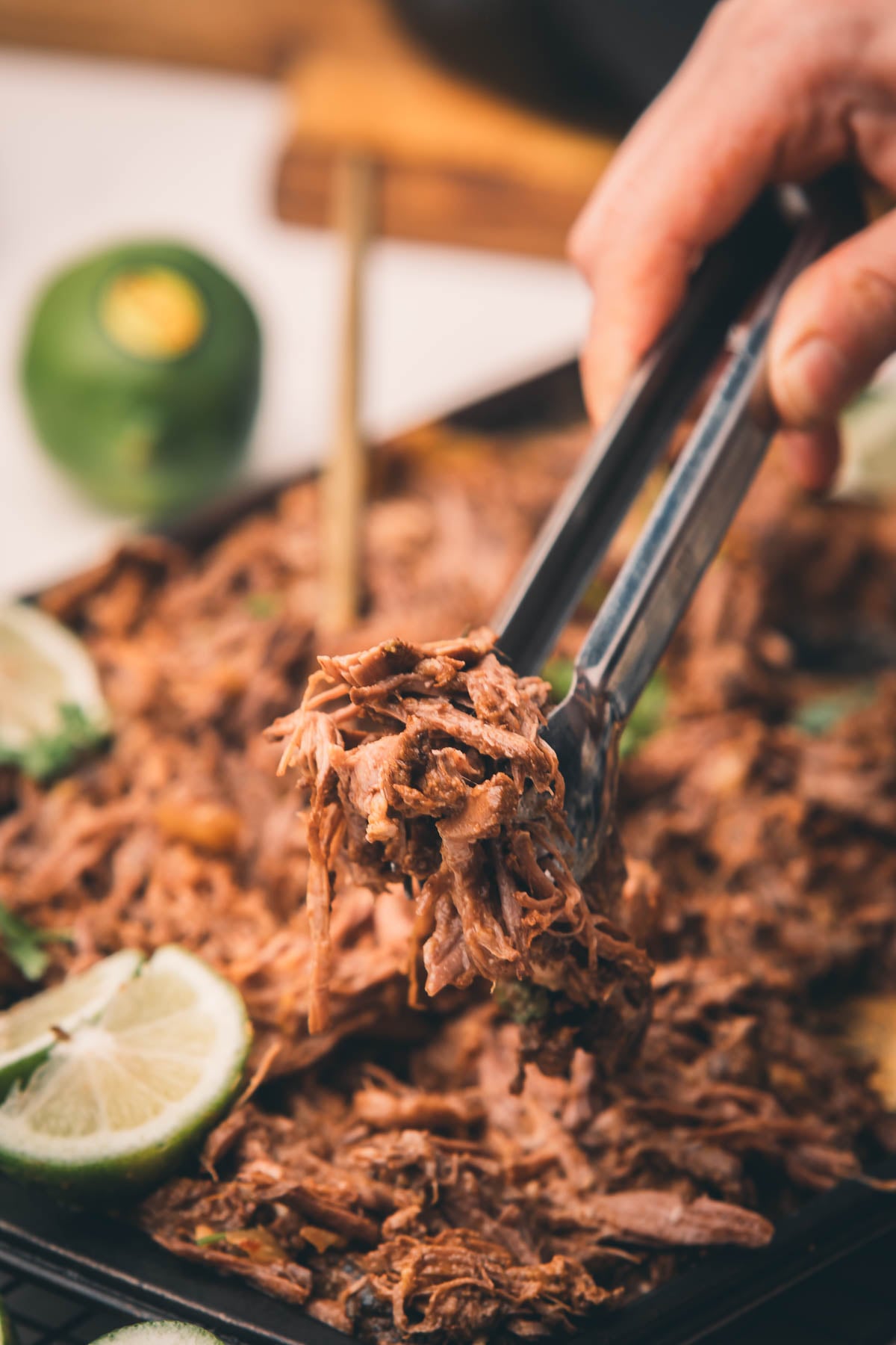 A hand uses tongs to pick up shredded beef from a tray, with lime slices scattered around the meat.