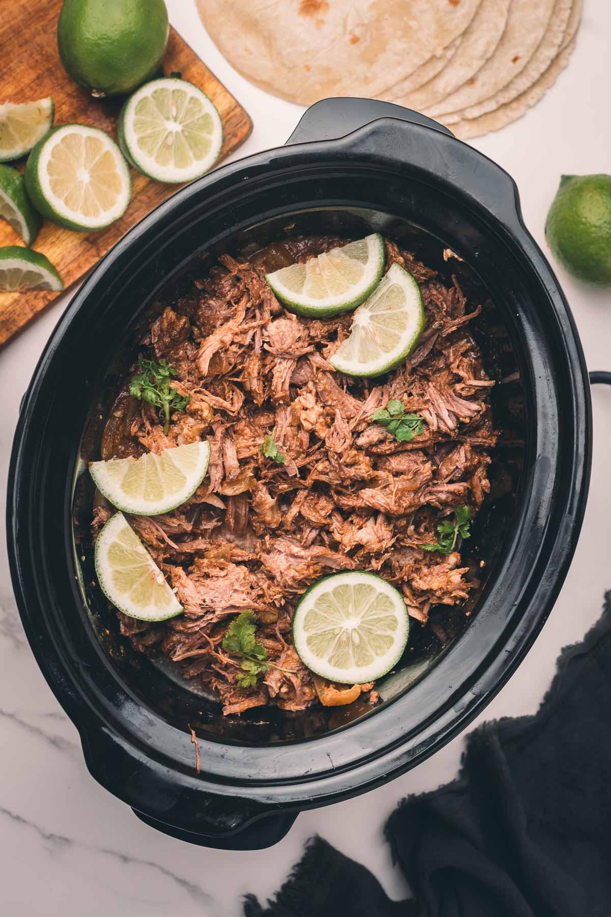 Shredded Mexican beef in a slow cooker topped with lime slices, with tortillas, halved limes, and lime slices on a wooden board nearby.