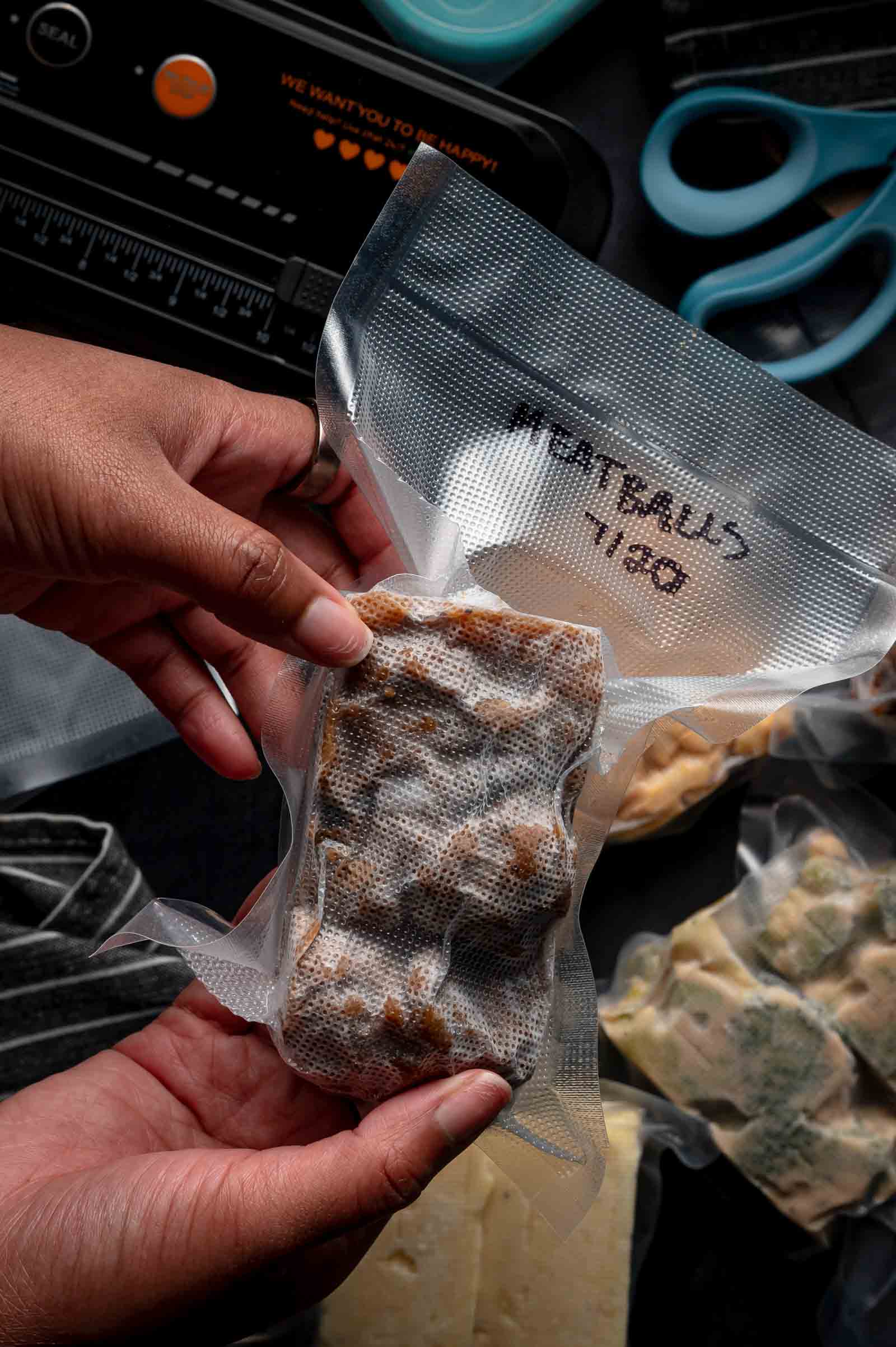 A person holds a vacuum-sealed bag labeled "Meatballs 7/20" next to a vacuum sealer, scissors, and other sealed food items on a dark surface.