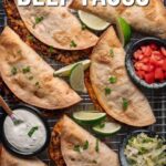 Five baked crispy beef tacos are arranged on a wire rack with lime wedges, chopped tomatoes, shredded lettuce, and a bowl of sauce on the side.