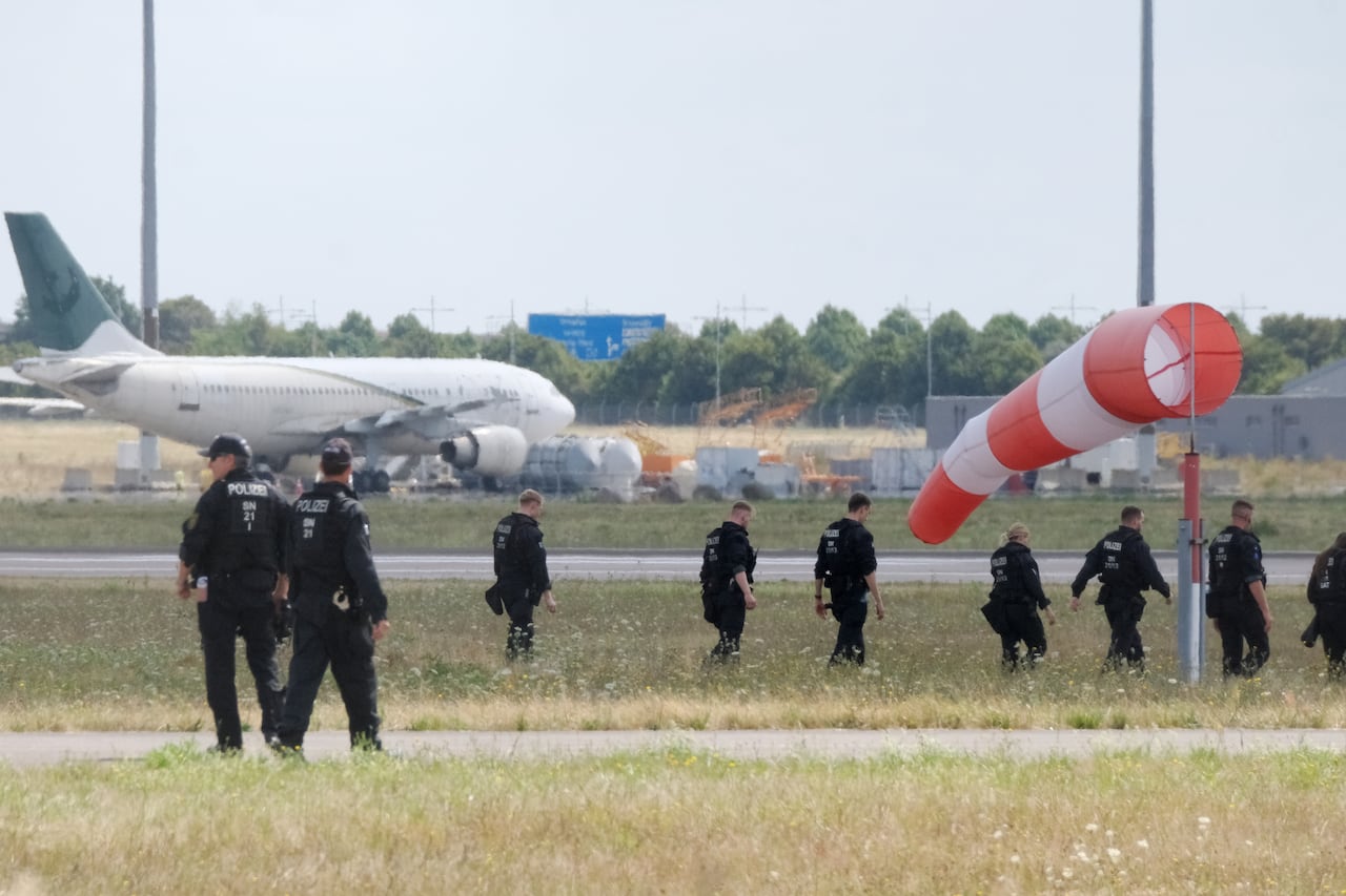 From a distance, about a dozen people in dark uniforms walk in a field that is located not far from where a couple of airplanes are shown.