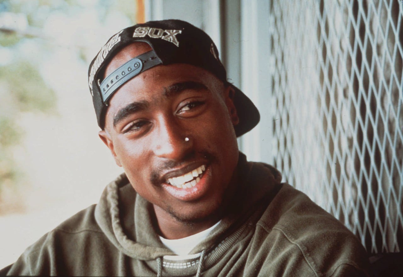 A smiling man in a backwards baseball cap and green hoodie in front of a fence.