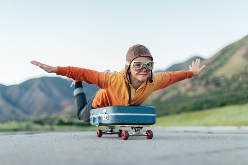 A young boy wearing flying goggles and flight cap has packed his suitcase and has placed it on a skateboard. He is ready to fly away with arms outstretched to the destination of his dreams. 